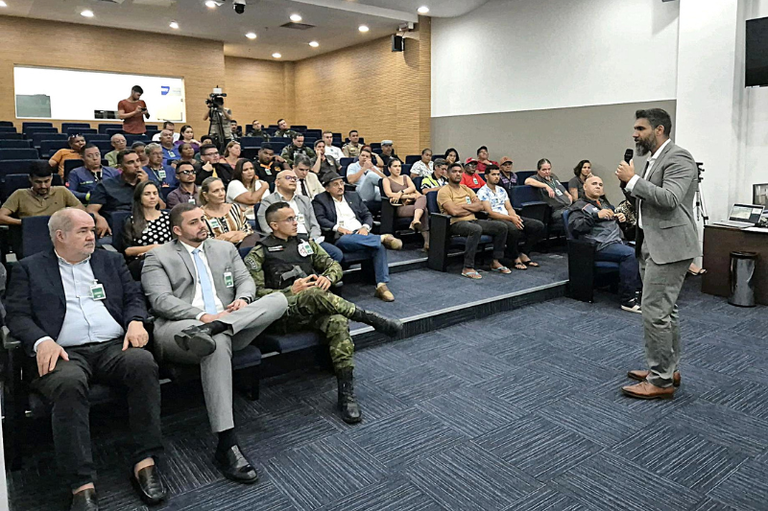 Um homem vestido com terno cinza está em pé, falando ao microfone diante de uma plateia reunida em auditório.