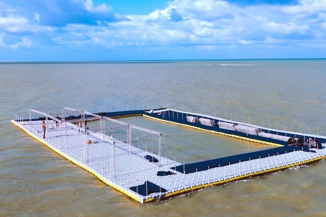 Fotografia de uma estrutura retangular flutuante sobre o mar, nas cores branca e preta, com operários trabalhando na montagem.