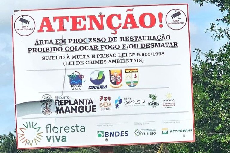 A imagem mostra uma placa grande de aviso ambiental instalada em uma área verde, com árvores ao fundo e céu parcialmente azul. A placa tem moldura vermelha e fundo branco, com o título em letras grandes e vermelhas: ATENÇÃO!
O texto informa que o local está em processo de restauração ambiental, proibindo colocar fogo ou desmatar, e menciona a Lei de Crimes Ambientais Lei nº 9.605/1998.

Na parte inferior, há vários logotipos coloridos de órgãos e projetos ambientais, como IBAMA, SUDEMA, BPAmb, Projeto Replanta Mangue, UFPB Campus IV, ICMBio, Floresta Viva, BNDES, FUNBIO e Petrobras.
Nos cantos superiores, há ícones circulares com uma imagem de câmera e o texto Área Monitorada, indicando vigilância.