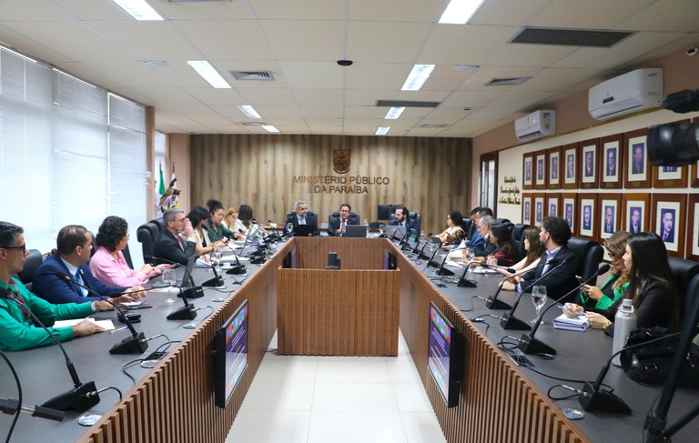 Em uma sala de reuniões ampla e formal, há uma mesa em formato de “U”, coberta por microfones, laptops e documentos. Diversas pessoas, vestidas de forma profissional, estão sentadas ao redor, participando de uma discussão.