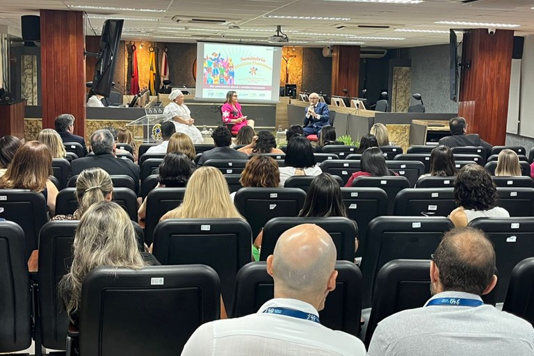 Auditório com paredes de madeira, cadeiras pretas ocupadas por público atento. No palco, três pessoas debatem diante de uma tela que exibe o título "Seminário Direitos Humanos: Por uma justiça plural e antirracista". Há bandeiras e um crucifixo ao fundo, indicando ambiente institucional.