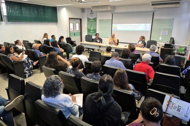 A imagem mostra um ambiente interno semelhante a um auditório. Diversas pessoas estão sentadas, voltadas para uma mesa comprida onde três pessoas participam de uma apresentação. Atrás deles, um projetor exibe o slide com o título "I Seminário de Reservas da Pesca no Estuário do rio Paraíba".