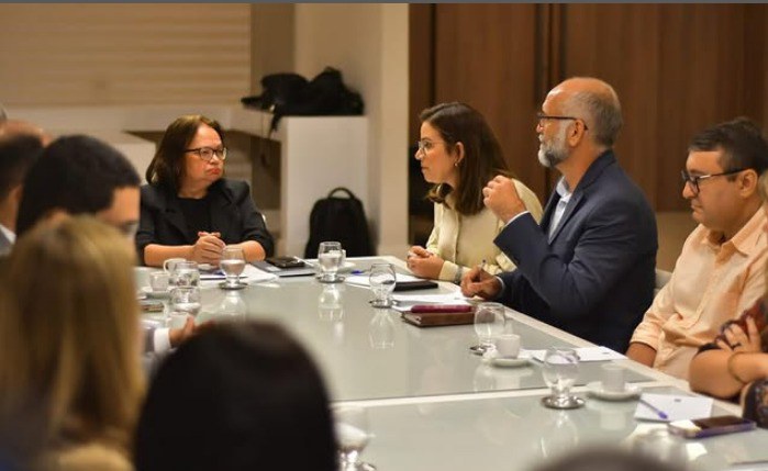 A imagem mostra uma reunião com varias pessoas sentadas ao redor de uma mesa retangular de vidro.