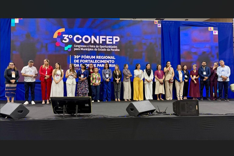 Um grupo de pessoas está alinhado no palco de um evento formal, diante de um telão azul que exibe o nome e as edições do congresso e fórum realizados no estado da Paraíba. Todos posam para a foto em ambiente iluminado e profissional.