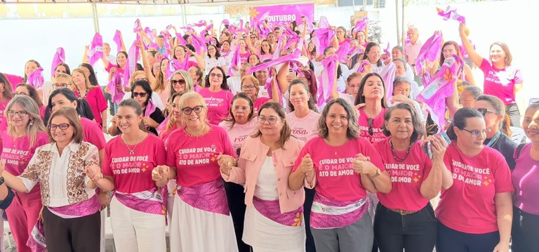 A imagem mostra um grande grupo de pessoas reunidas em um evento, a maioria vestindo camisetas cor-de-rosa e segurando lenços ou fitas da mesma cor. Na frente, algumas camisetas trazem a frase “Cuidar de você é o maior ato de amor”. Ao fundo, há um banner com a palavra “Outubro”, indicando que se trata de uma ação do movimento Outubro Rosa, voltado para a conscientização sobre o câncer de mama. O clima é de união e apoio coletivo.
