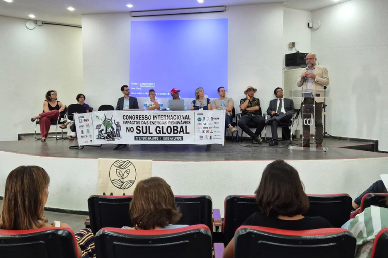 No palco de um auditório, há uma mesa comprida com vários palestrantes sentados. Um deles está em pé, falando ao público em um púlpito com a sigla CCJ. À frente da mesa, um banner anuncia um congresso internacional sobre impactos das energias renováveis no Sul Global. O público está sentado em cadeiras de teatro, acompanhando a apresentação.