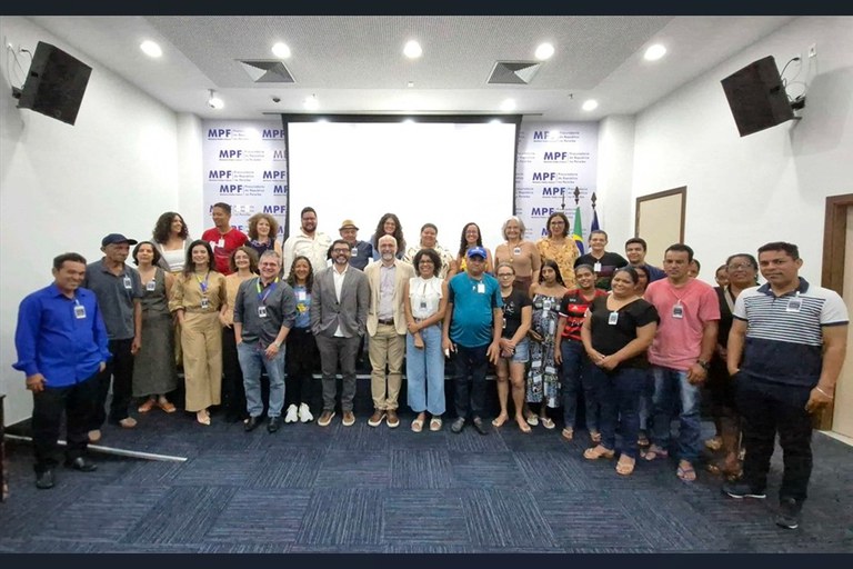 A imagem mostra um grupo de cerca de trinta pessoas reunidas em uma sala de conferências. Elas estão dispostas em duas fileiras, algumas usando crachás de identificação. Ao fundo, há uma tela de projeção branca e uma parede com o logotipo repetido do MPF (Ministério Público Federal).