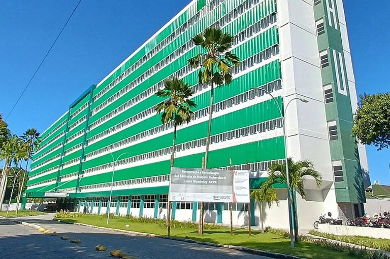 Fachada de um grande edifício hospitalar nas cores verde e branco, com palmeiras e uma placa informativa no gramado em primeiro plano, sob céu azul.