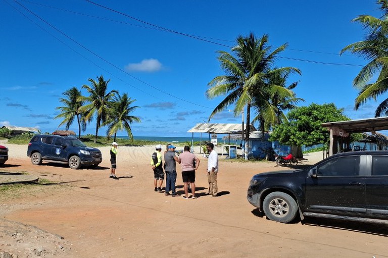 Foto mostra dois veículos estacionados e cinco pessoas conversando; ao fundo coqueiros e o mar