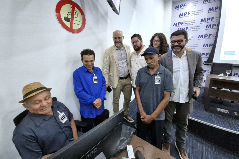 Um grupo de sete pessoas está reunido no auditório do MPF. No centro da cena, o agricultor Manoel Luiz,  presidente da Associação da Comunidade de Ponta de Gramame, está sentado diante de um computador, clicando para protocolar uma ação de reforma agrária na Justiça Federal. Ele observa atentamente a tela. Ao seu redor, pessoas acompanham o momento com expressões de atenção e seriedade. Ao fundo, há um painel institucional do MPF e um telão de projeção. A atmosfera é formal e registra um ato oficial importante para a comunidade.