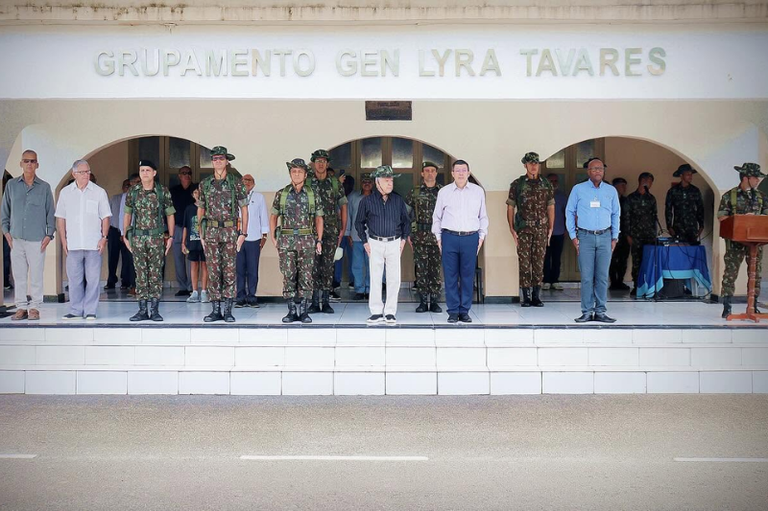 Militares em farda camuflada e civis enfileirados em posição de sentido sobre uma plataforma elevada. Ao fundo, a fachada de um prédio com arcos exibe a inscrição "GRUPAMENTO GEN LYRA TAVARES".