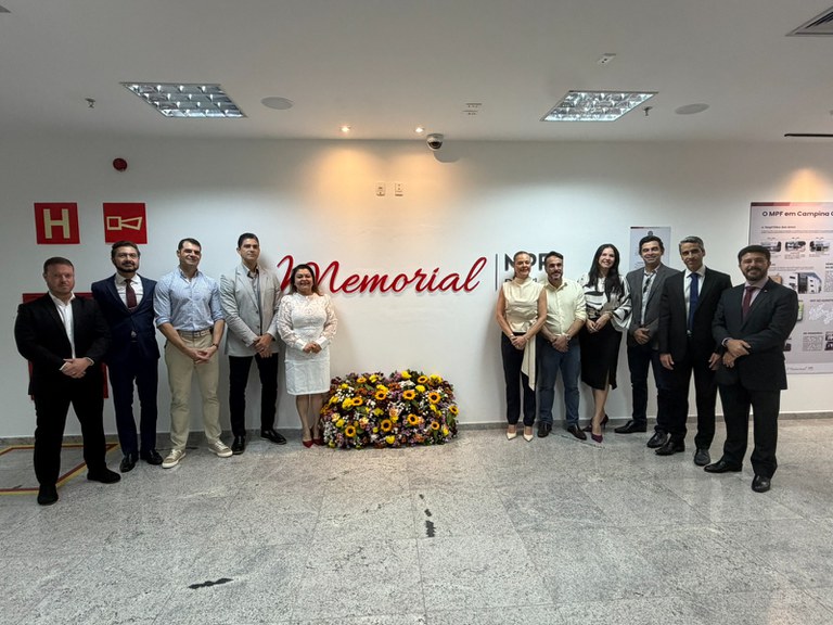 Um grupo de pessoas está posicionado lado a lado em um ambiente interno. Ao fundo, na parede, há o texto em vermelho Memorial MPF e, abaixo dele, um grande arranjo de flores coloridas.