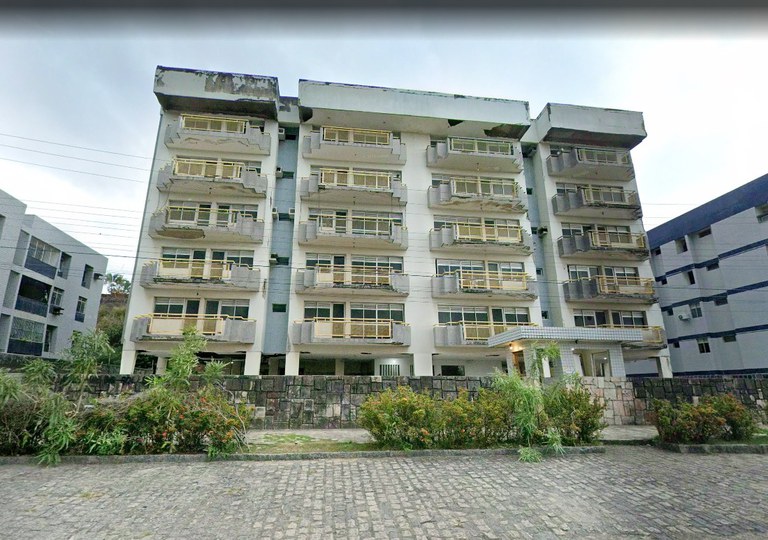 A imagem mostra um prédio residencial de seis andares, pintado em tons de branco e cinza. Cada andar possui varandas com grades claras, e a fachada apresenta sinais de desgaste próximo ao telhado