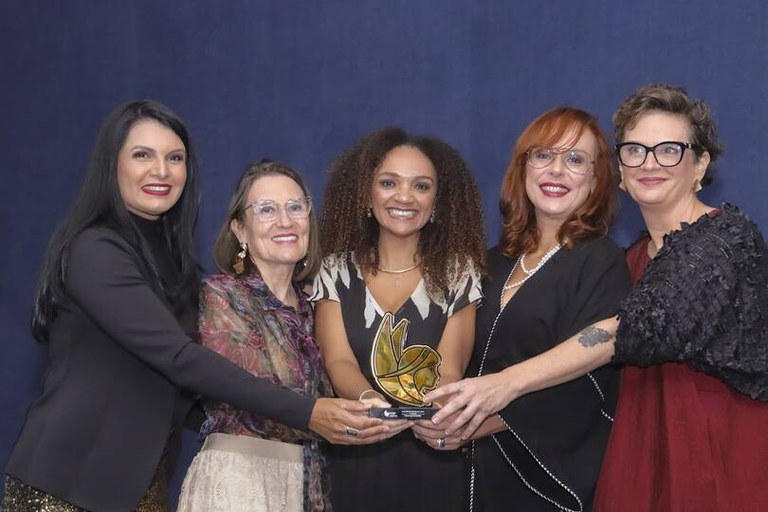 Cinco mulheres posam lado a lado diante de um fundo azul-escuro. Todas sorriem para a câmera enquanto seguram juntas um troféu dourado em formato estilizado. Elas vestem roupas sociais em tons de preto, vinho, bege e estampado, em um registro feito durante cerimônia de premiação.