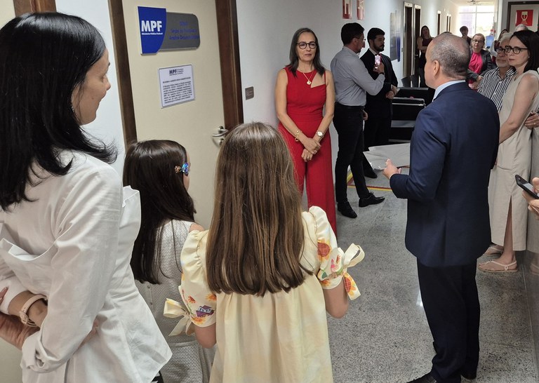 A imagem mostra um grupo de pessoas reunidas em um corredor bem iluminado. Há uma porta com uma placa identificando MPF e o nome do setor do servidor homenageado.