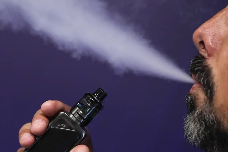 Homem com barba exala vapor pela boca enquanto segura um cigarro eletrônico preto, em frente a um fundo roxo.