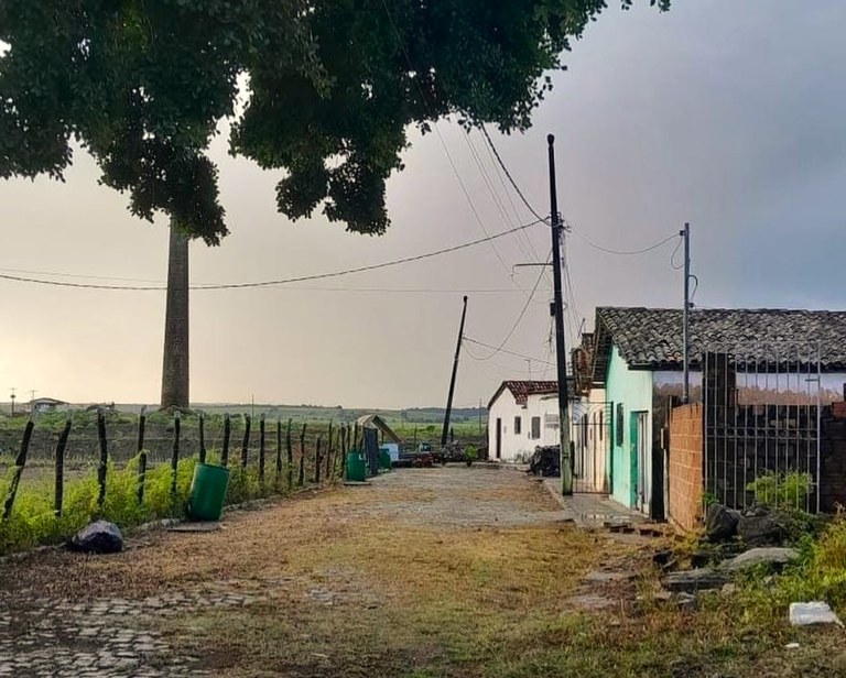 Imagem mostra uma rua de terra batida e paralelepípedo em uma área rural, com casas simples à direita e uma cerca de madeira à esquerda. Ao fundo, vê-se uma antiga chaminé alta, remanescente de engenho. O céu está nublado e a paisagem ao redor é de campo aberto.