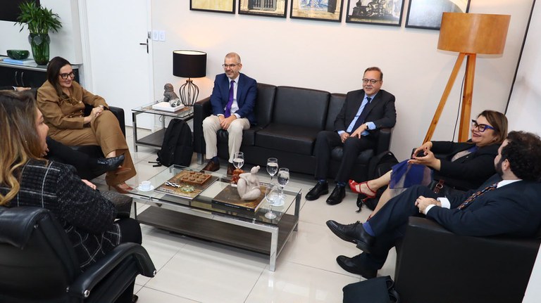 A imagem mostra sete pessoas reunidas em uma sala de reunião moderna. Elas estão sentadas em sofás e poltronas pretas dispostas em torno de uma mesa de centro de vidro, que tem objetos decorativos, copos com água e uma escultura.