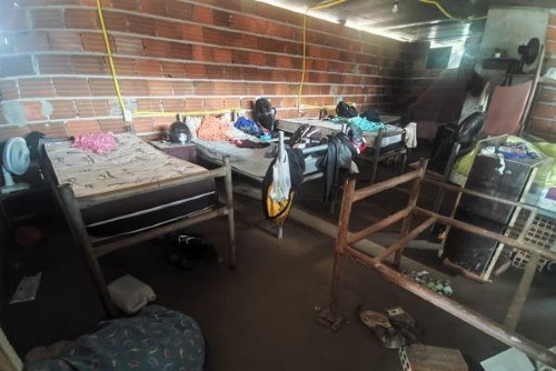 Foto de um lugar em construção onde se vê camas, colchões, ventiladores e roupas espalhadas.