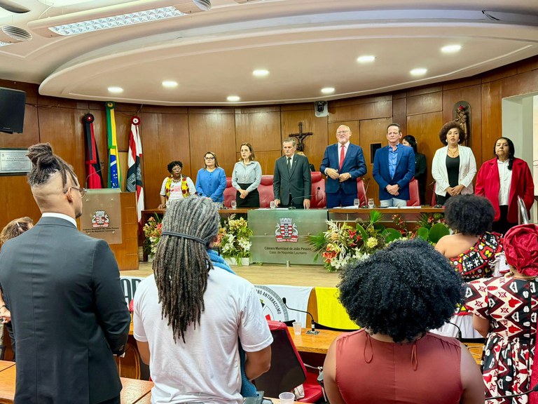 A imagem mostra uma cerimônia formal no plenário da Câmara Municipal de João Pessoa. Ao fundo, dez pessoas estão em pé atrás de uma longa bancada decorada com arranjos florais.