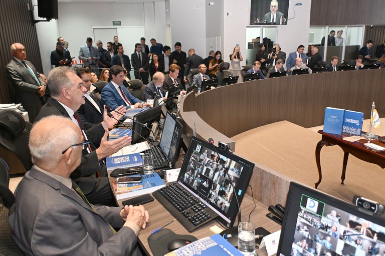 A imagem mostra uma reunião formal em um ambiente moderno. Pessoas estão sentadas em torno de uma mesa curva equipada com computadores e microfones, participando de uma discussão. Nos monitores, aparecem participantes conectados por videoconferência. Ao fundo, há observadores em pé, alguns registrando fotos, reforçando o caráter oficial e institucional do encontro.