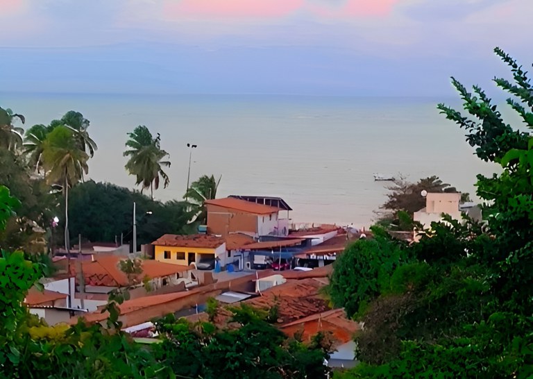 A imagem mostra uma vista panorâmica da comunidade quilombola pesqueira da praia da Penha, em João Pessoa. No primeiro plano, há casas simples com telhados de barro e paredes coloridas, cercadas por vegetação verde intensa. Algumas palmeiras altas se destacam, balançando suavemente ao vento. Ao fundo, o mar aparece calmo e claro, refletindo tons suaves de azul e rosa do céu, que indicam o amanhecer ou entardecer. O cenário transmite tranquilidade e um forte vínculo entre natureza e vida comunitária à beira-mar.