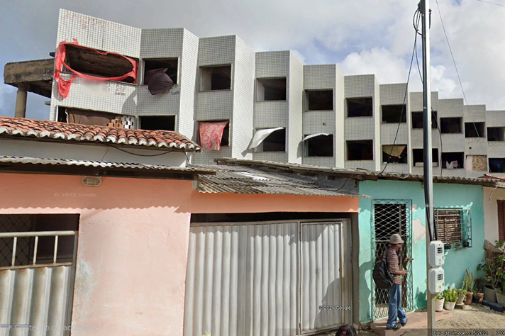 A imagem mostra uma rua com construções contrastantes. Em primeiro plano, há duas casas térreas simples: uma pintada de rosa claro com telhado de cerâmica, e outra azul com portão metálico e fachada desgastada. Uma pessoa de chapéu e mochila está em frente à casa azul, aparentemente mexendo no celular. Ao fundo, destaca-se um prédio inacabado de vários andares, com fachada revestida de pastilhas brancas e janelas quadradas sem vidros, algumas cobertas por panos ou cortinas improvisadas.