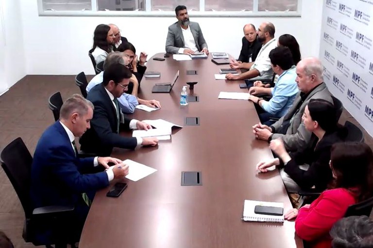 Foto mostra a mesa de reuniões com 14 pessoas sentadas ao redor, em cima da mesa, materiais como papéis, computador, canetas e celulares; na parede do lado direito um banner do MPF entre a porta e as janelas