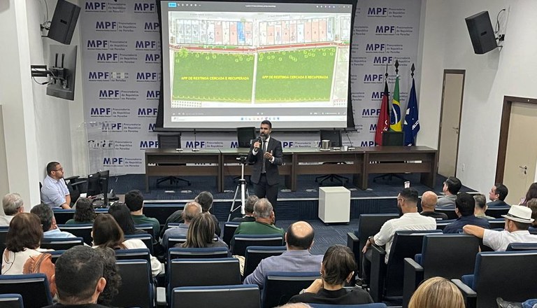 A imagem mostra uma sala de conferência formal, com várias pessoas sentadas e voltadas para um palestrante. O palestrante está em pé, usando microfone, diante de uma tela de projeção grande