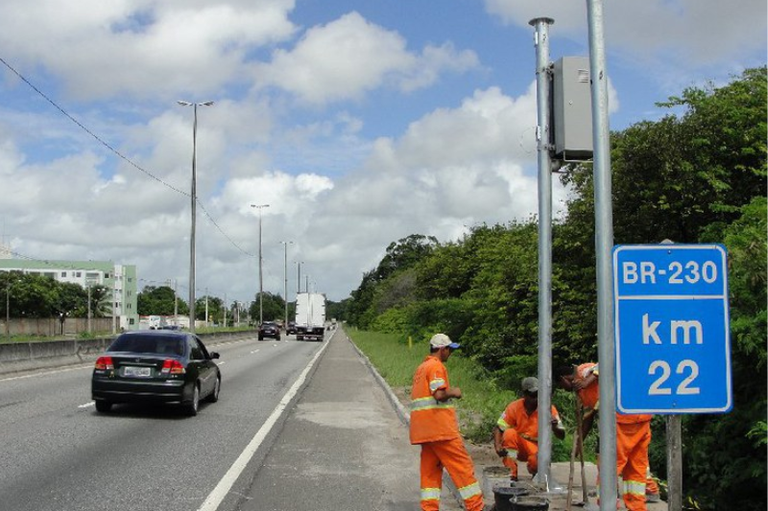 Homens usando uniformes laranja de manutenção trabalham na lateral de uma rodovia movimentada, a BR-230, no quilômetro 22, conforme sinalização visível no poste ao lado de um equipamento. Veículos, incluindo um caminhão, circulam na pista.