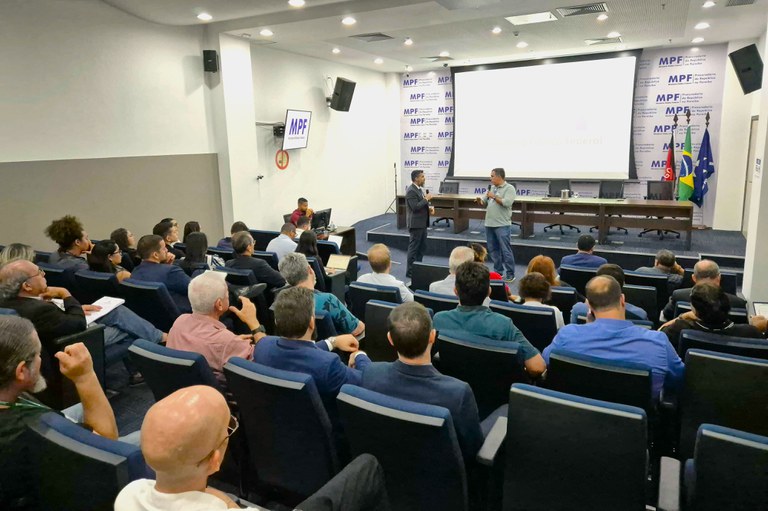 A imagem mostra um auditório amplo, com várias pessoas sentadas e voltadas para o palco. No palco, dois palestrantes estão em pé diante de uma tela de projeção grande. O fundo do palco traz repetidamente o logotipo MPF e Ministério Público Federal.