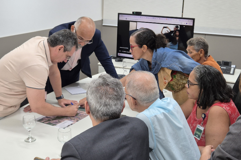 Foto mostra um grupo de pessoas reunidas ao redor de uma mesa em uma sala de reuniões, analisando e discutindo mapas espalhados sobre a superfície. Dois indivíduos estão de pé, apontando para os mapas, enquanto os demais observam e participam da conversa. Há um monitor exibindo uma videochamada ou apresentação, refletindo a mesma cena na mesa
