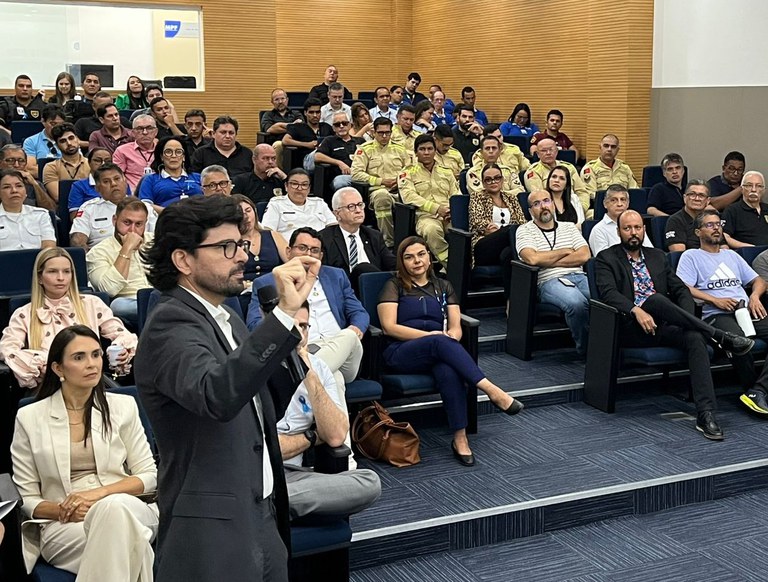 Um homem de terno escuro, em primeiro plano, fala em um auditório lotado, gesticulando com a mão direita. O público, composto por homens e mulheres com uniformes e roupas casuais, assiste atentamente.