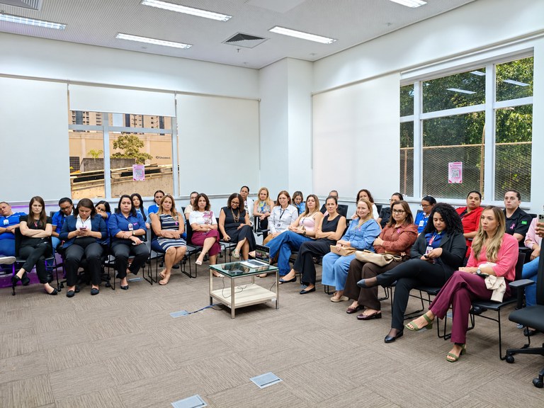 Sala de reunião ampla e bem iluminada, com paredes claras, janelas grandes e piso acarpetado. Cerca de trinta pessoas estão sentadas em cadeiras organizadas em semicírculo, voltadas para o centro da sala.