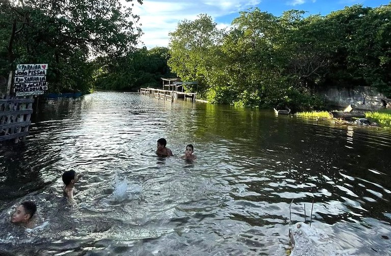 A imagem retrata um rio cercado por vegetação densa e árvores verdes. O céu azul, com nuvens brancas espalhadas em formações onduladas, reflete-se na água. Algumas crianças estão nadando e brincando no rio, criando pequenos respingos. À esquerda, há uma placa escrita à mão indicando “João Pessoa” e Rio Sanhauá. Mais ao fundo, vê-se uma pequena estrutura de madeira à beira do rio. No canto superior direito, parte do telhado de uma construção aparece na imagem. A cena transmite um clima de tranquilidade e lazer em meio à natureza.
