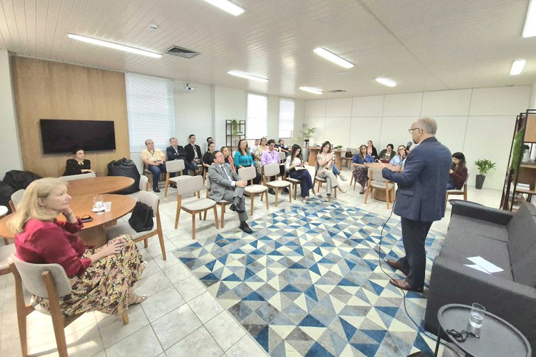 Em uma sala de conferências, um homem em pé, no primeiro plano, segura um microfone e fala para o público. À frente dele, várias pessoas estão sentadas em cadeiras dispostas em fileiras, prestando atenção. O ambiente é bem iluminado, com piso claro e um tapete de padrão geométrico em azul e cinza.