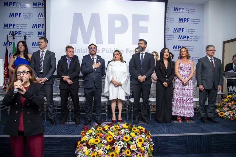 A imagem mostra um evento solene realizado no Ministério Público Federal da Paraíba. No palco, várias pessoas estão alinhadas, vestindo trajes formais, diante de um grande painel com o logotipo do MPF. À frente do palco, há arranjos florais coloridos. Uma mulher em primeiro plano aparece cantando. O ambiente transmite a atmosfera de uma cerimônia oficial, com destaque para a formalidade e a representatividade institucional.