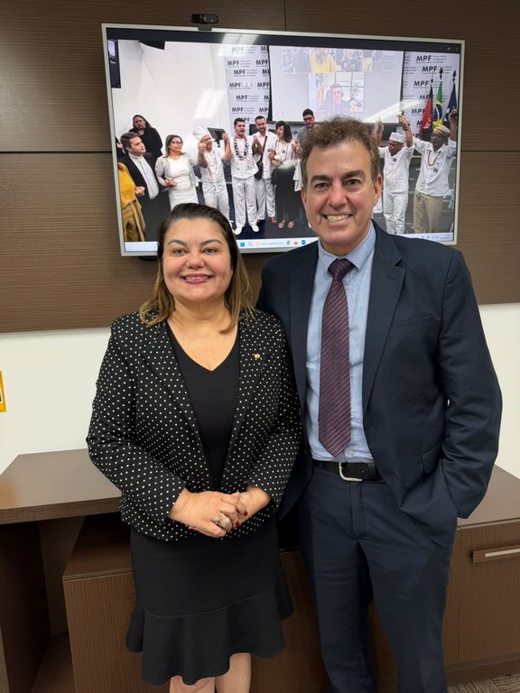 Duas pessoas posam lado a lado em um ambiente institucional. À frente de uma tela de videoconferência exibindo um grupo de pessoas vestidas de branco, uma mulher usa blazer preto com detalhes em branco e vestido preto, enquanto um homem veste terno azul-marinho com gravata vinho. Ambos sorriem para a câmera. Ao fundo, aparecem elementos visuais do Ministério Público Federal.