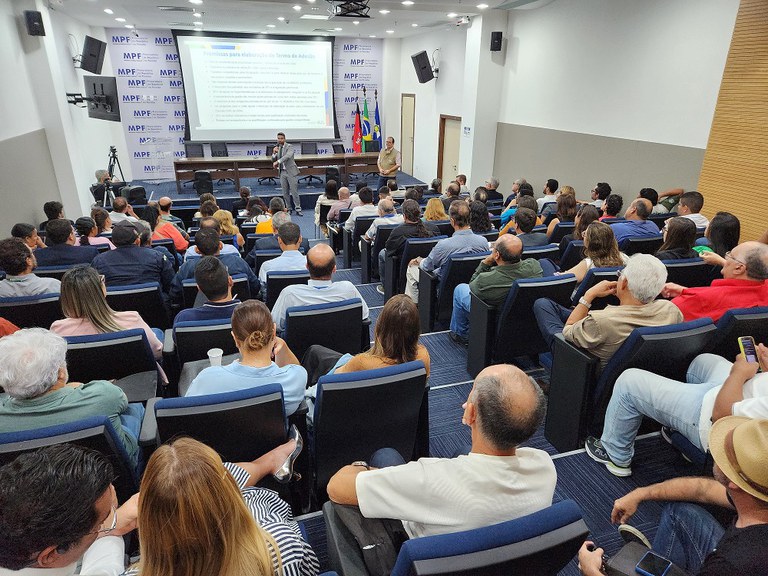 A imagem mostra um auditório lotado, com diversas pessoas sentadas em cadeiras azuis, assistindo a uma apresentação. No palco, um homem de terno segura um microfone e fala ao público, enquanto outro homem, de camisa bege, está ao seu lado. No fundo, há uma tela de projeção exibindo um slide com texto e os logotipos do Ministério Público Federal repetidos no painel atrás dos palestrantes. Algumas pessoas na plateia estão usando celulares, enquanto outras observam atentamente a apresentação. O ambiente é iluminado e tem um design moderno, com paredes claras e detalhes em madeira.