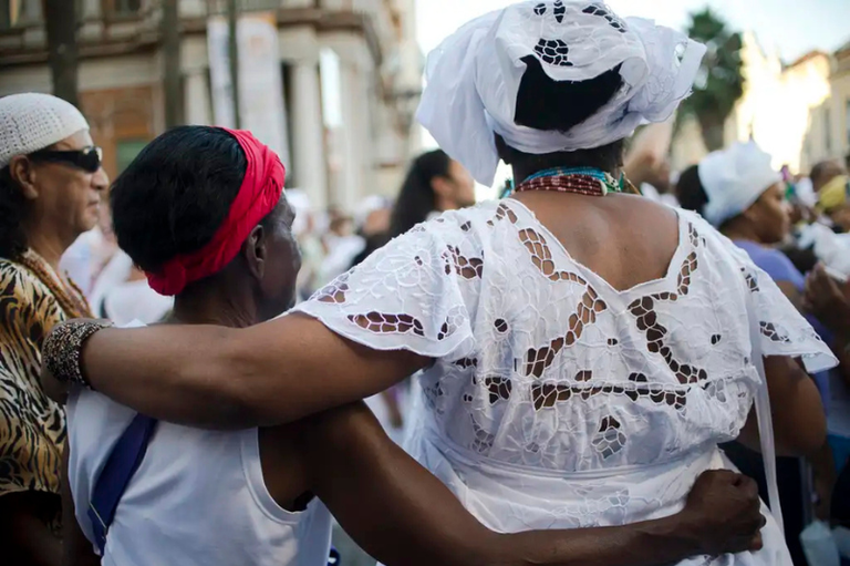 Foto de duas pessoas de religiões de matriz africana, vestidas de branco e turbantes (uma delas de costas), abraçadas durante um evento de rua.