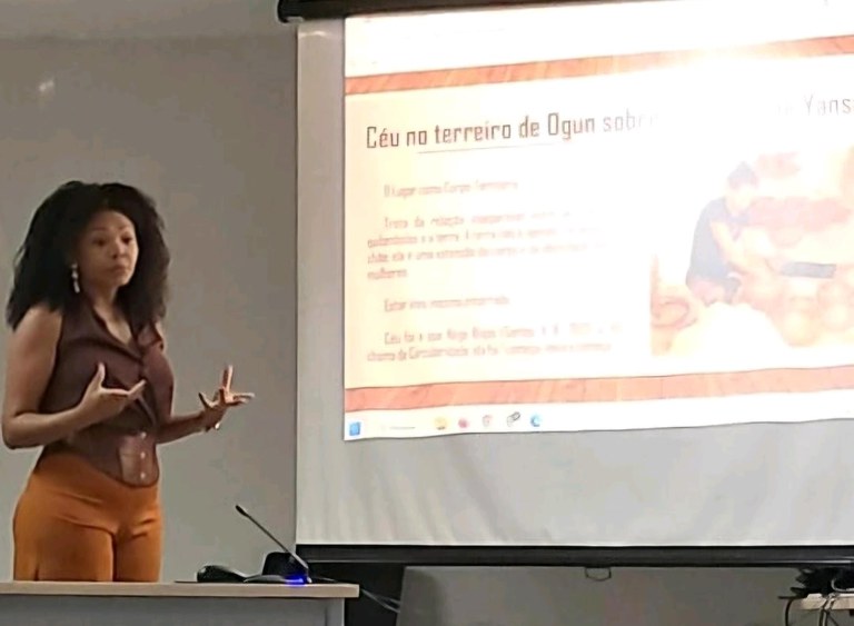 A imagem mostra uma mulher negra em pé, diante de uma tela projetada em um ambiente de sala de aula ou conferência. Ela está gesticulando com as mãos, em postura ativa de explicação, indicando que conduz uma apresentação.
