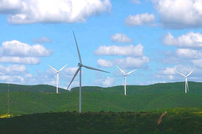 A imagem mostra um campo de turbinas eólicas sobre colinas verdes.