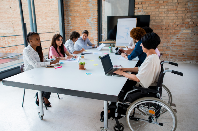 Grupo diverso de profissionais sorridentes em um ambiente de escritório moderno, com um homem em cadeira de rodas em destaque no primeiro plano.