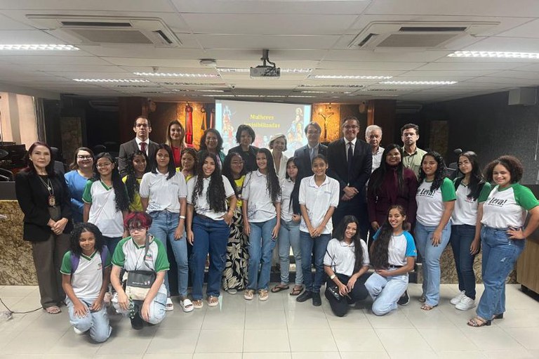 Fotografia colorida de um grupo de aproximadamente 30 pessoas posando para uma foto em um auditório. A maioria são jovens estudantes, com alguns adultos e autoridades em destaque, todos em pé ou agachados. No fundo, há uma tela de projeção com o texto "Mulheres Invisibilizadas". O ambiente parece ser um evento oficial ou uma palestra.