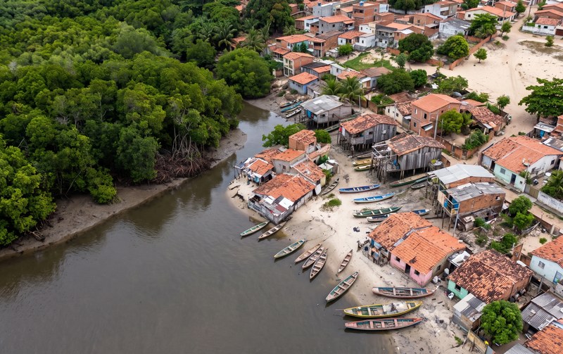 Vista aérea de uma comunidade ribeirinha. À esquerda, uma extensa área de vegetação densa e verde acompanha um braço de rio de águas escuras. À direita, casas simples com telhados de cerâmica avermelhados se distribuem ao longo da margem arenosa. Diversas canoas e pequenos barcos estão atracados na beira do rio. Algumas construções de madeira aparecem elevadas sobre estacas próximas à água. Ao fundo, mais casas e ruas de terra compõem o cenário, cercado por árvores e áreas verdes.