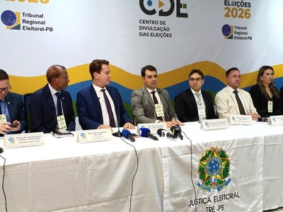 Em uma sala de coletiva de imprensa, sete pessoas estão sentadas atrás de uma mesa longa coberta por uma toalha branca com o brasão da Justiça Eleitoral. No centro da mesa, há vários microfones voltados para os participantes. Ao fundo, um painel exibe os logotipos do Tribunal Regional Eleitoral da Paraíba, do Centro de Divulgação das Eleições e a inscrição Eleições 2026. Os participantes usam trajes formais e estão posicionados lado a lado, voltados para o público.