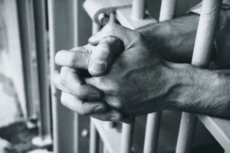 Imagem de uma pessoa dentro de uma cela e as mãos cruzadas