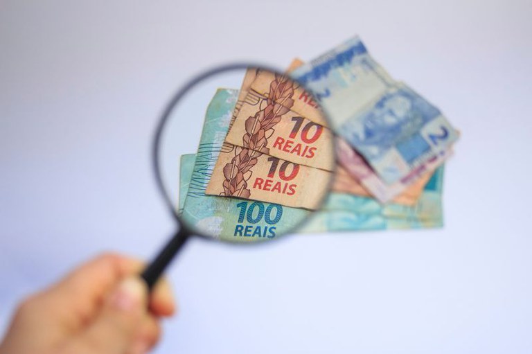 Mão segurando uma lupa sobre um monte de notas de dinheiro brasileiro (reais) sobre fundo claro.