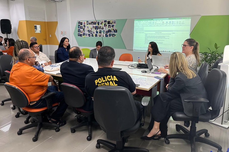 A imagem mostra um grupo de nove pessoas reunidas em torno de uma mesa retangular em uma sala bem iluminada, com cadeiras de escritório e uma tela projetando uma planilha visível na parede. Algumas pessoas estão olhando para a tela, enquanto outras parecem estar conversando ou olhando para os papéis sobre a mesa. Há também um quadro branco com anotações visível na mesa.