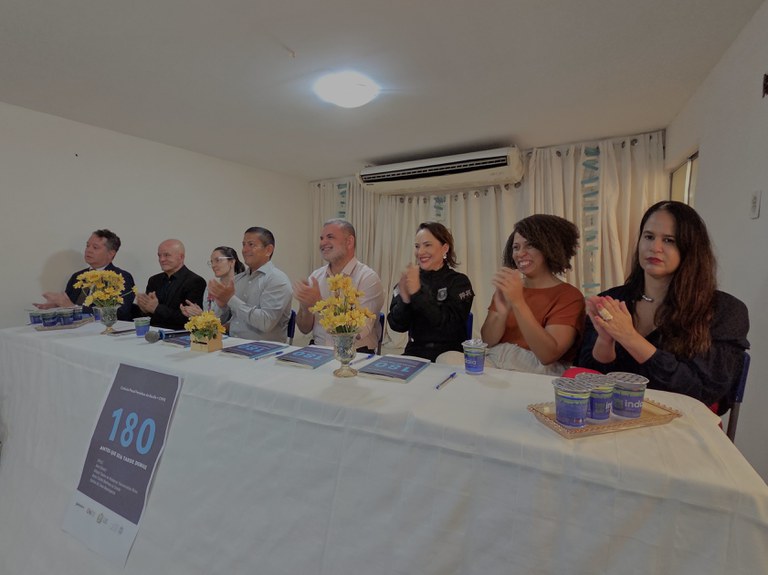 A imagem mostra sete representantes de órgãos sentados em uma mesa, a maioria aplaudindo diante de arranjos de flores amarelas e copos d'água. Na frente da mesa coberta com toalha branca, destaca-se um cartaz impresso com o número "180".